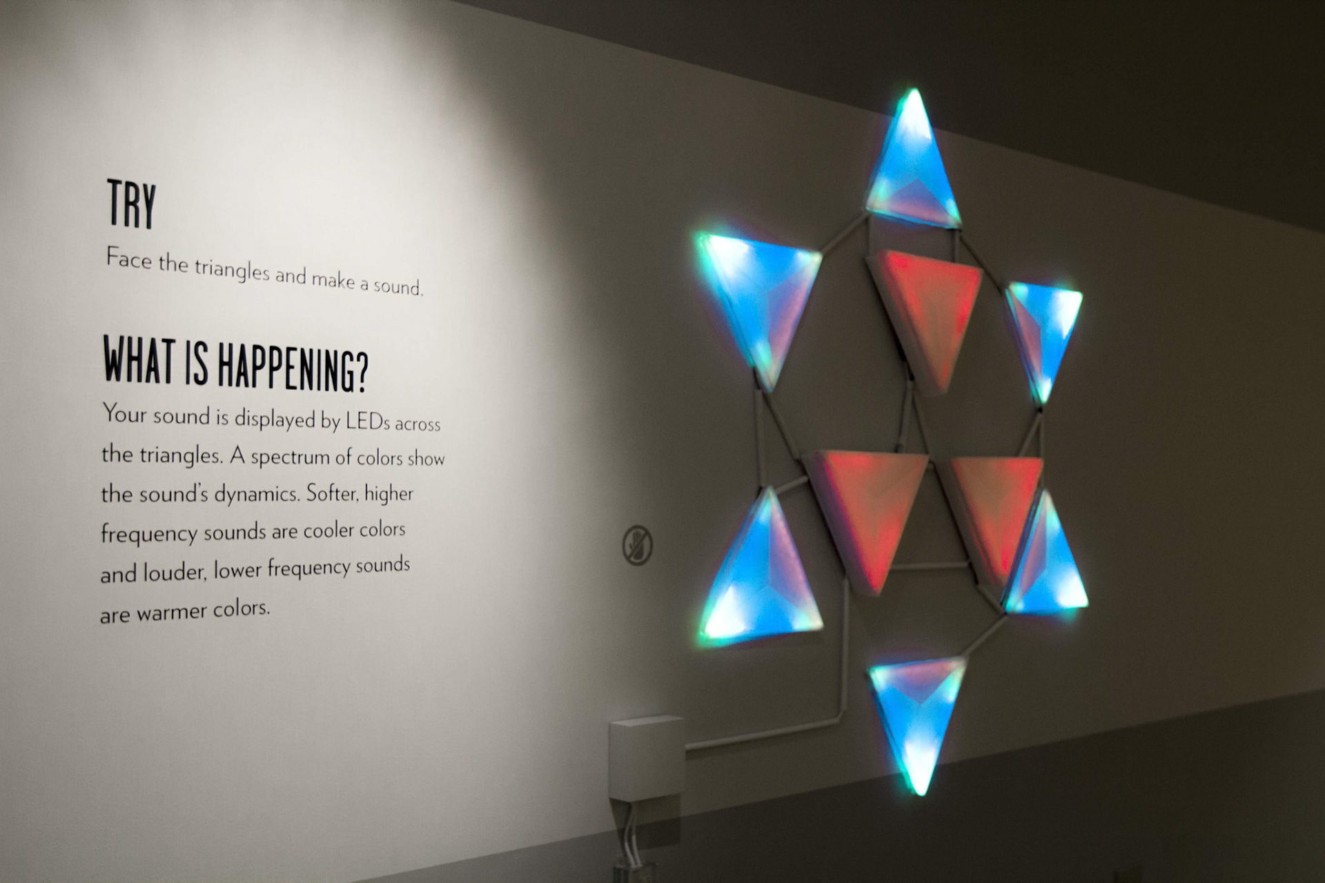 The nine-triangle star lit on a pale museum wall, the six outer triangles glowing blue and cyan and the three inner ones red, beside wall text reading “TRY: Face the triangles and make a sound”