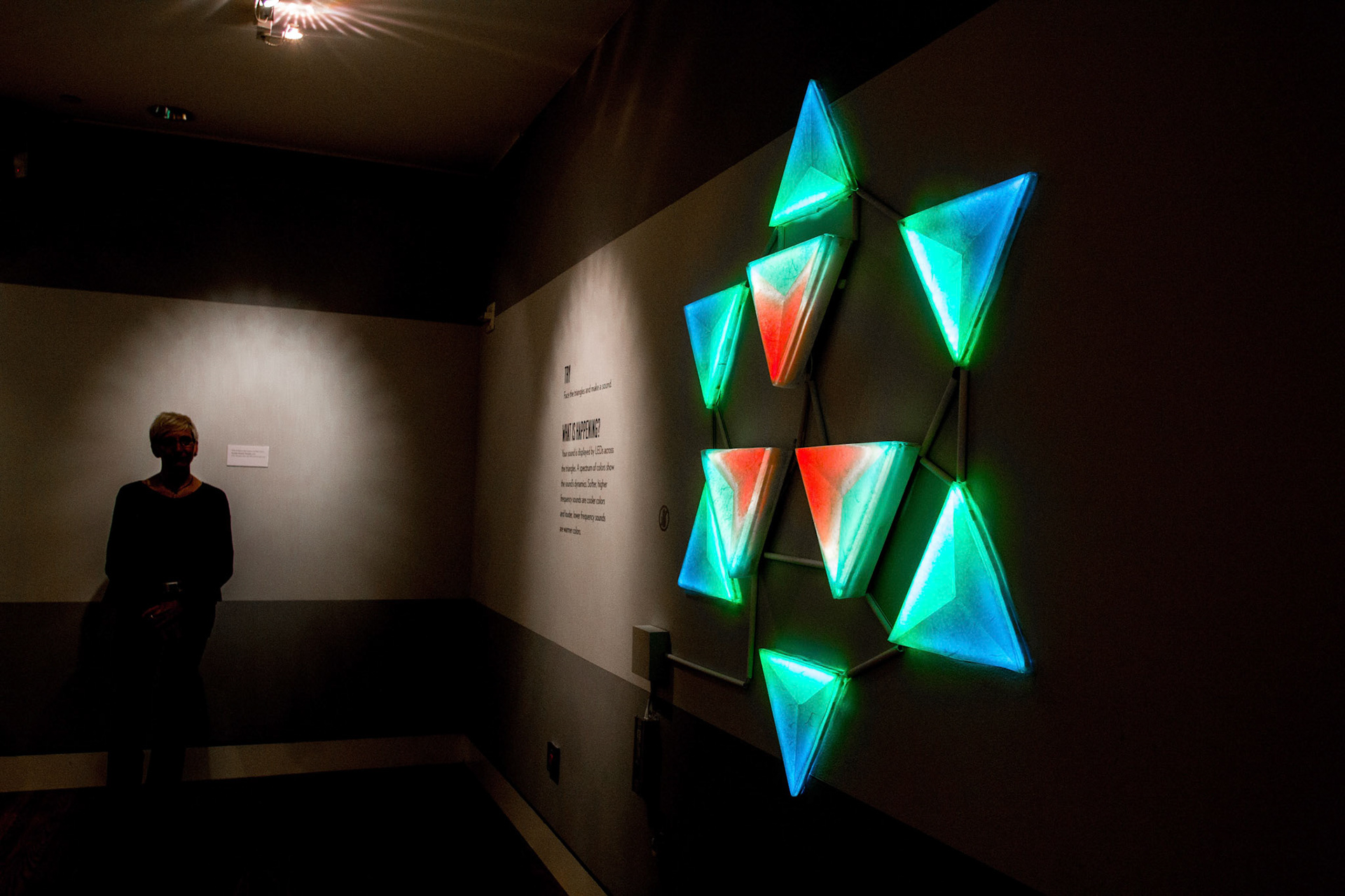 A visitor stands in a darkened gallery beside Cymatic Star, a wall-mounted cluster of triangular panels glowing green, blue and red