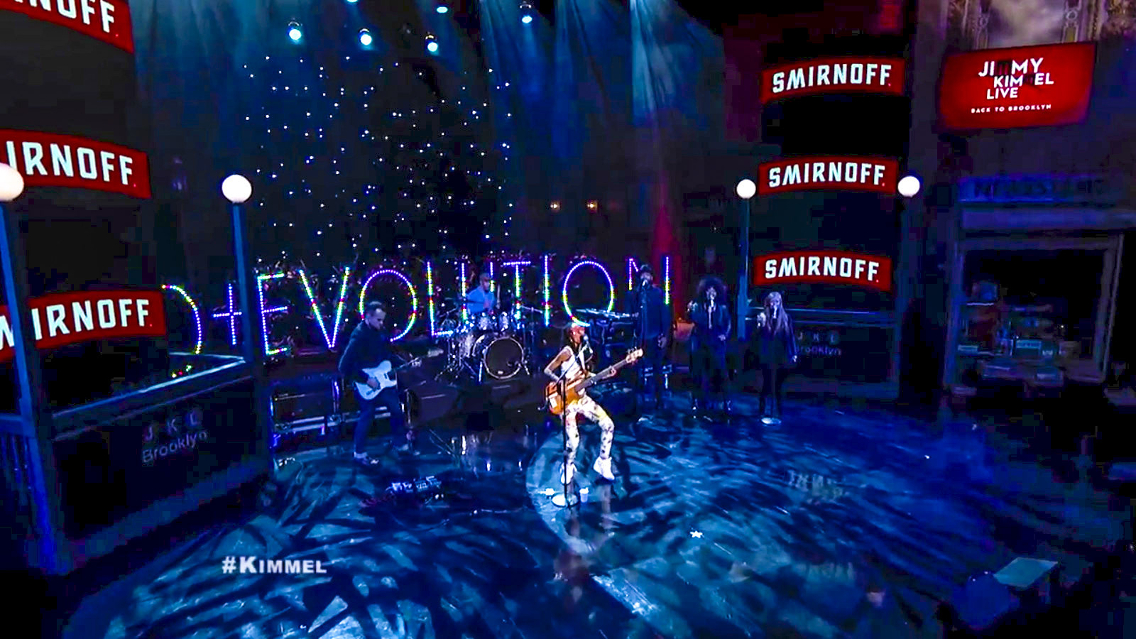 Wide view of the Jimmy Kimmel Live Brooklyn set: the full D+EVOLUTION LED sign spans the stage behind Spalding and her band, flanked by stacked Smirnoff signs, white streetlamp globes and a pinpoint starfield backdrop