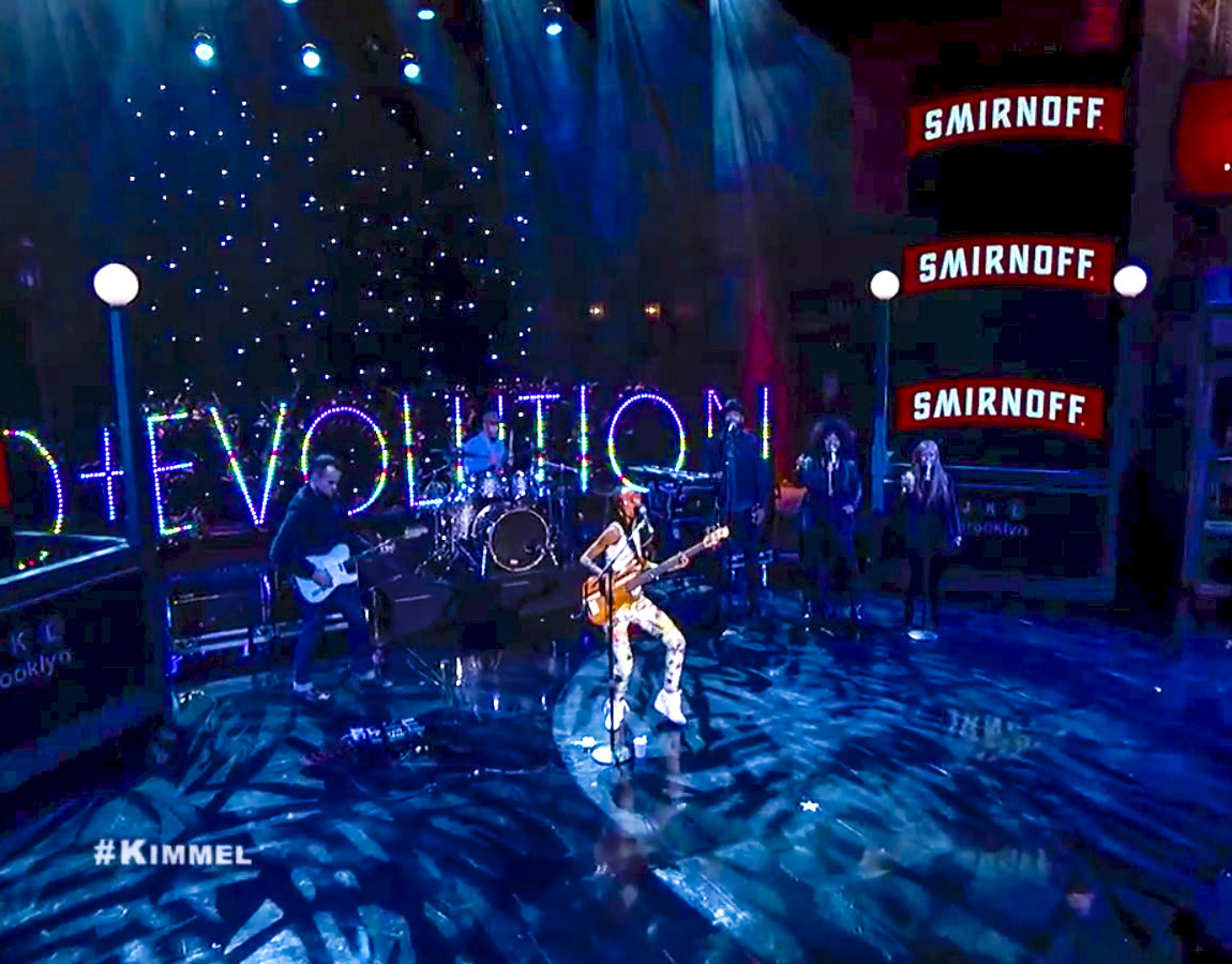 Esperanza Spalding performing on the Jimmy Kimmel Live stage in front of a large illuminated EVOLUTION sign
