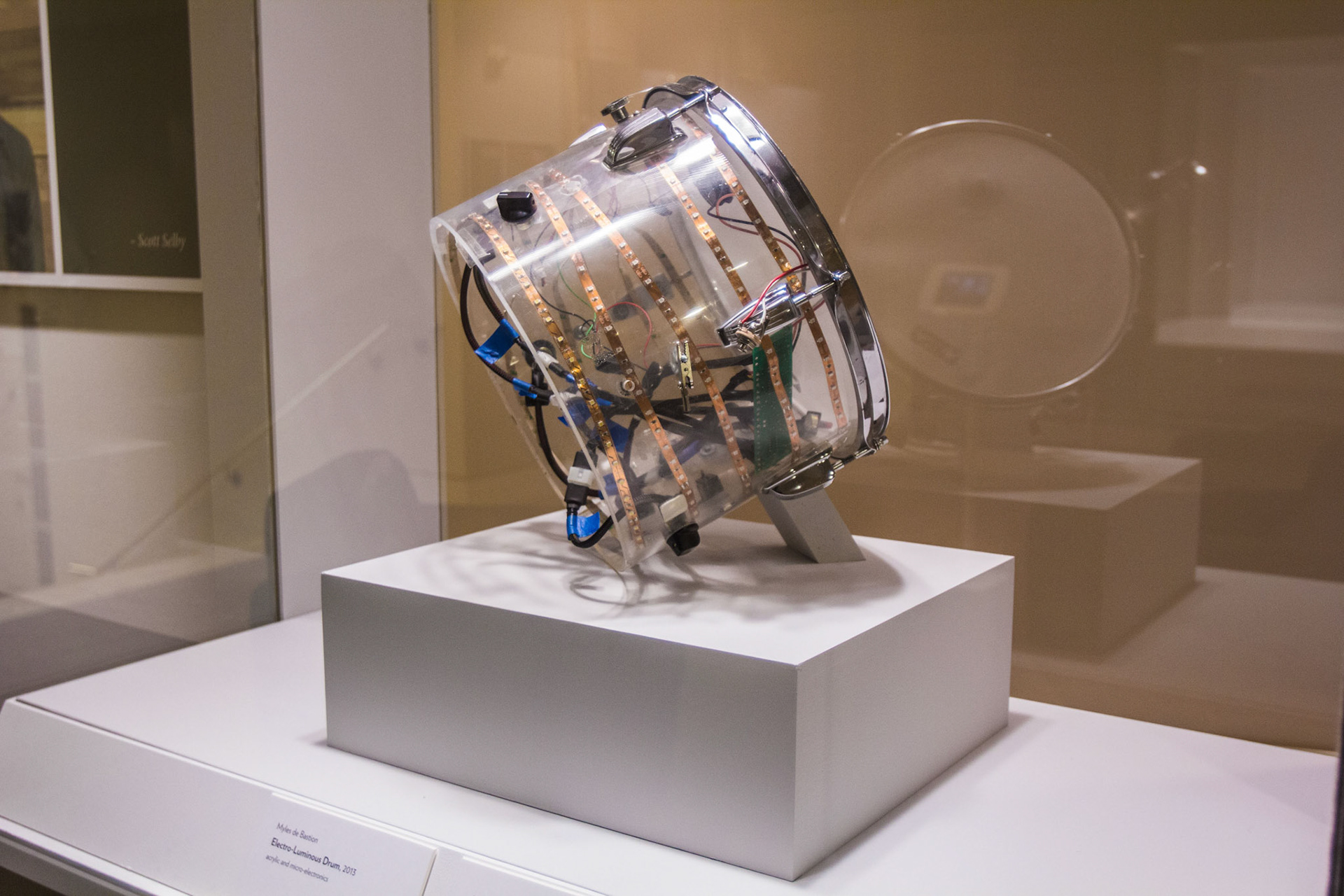 The Electro-Luminous Drum tilted on a stand inside its vitrine, a clear acrylic snare shell banded with copper LED strips and packed with colored wiring, its label reading Myles de Bastion, Electro-Luminous Drum, 2013