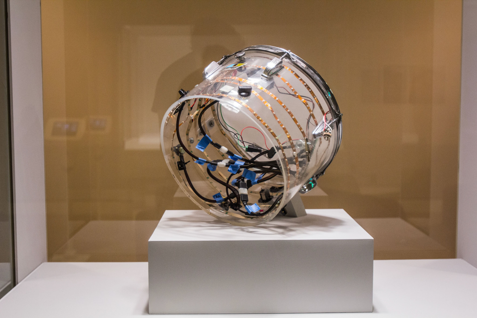 The Electro-Luminous Drum seen end-on against a warm tan vitrine wall, the open clear shell revealing vertical copper LED tape, black cables and strips of blue masking tape inside