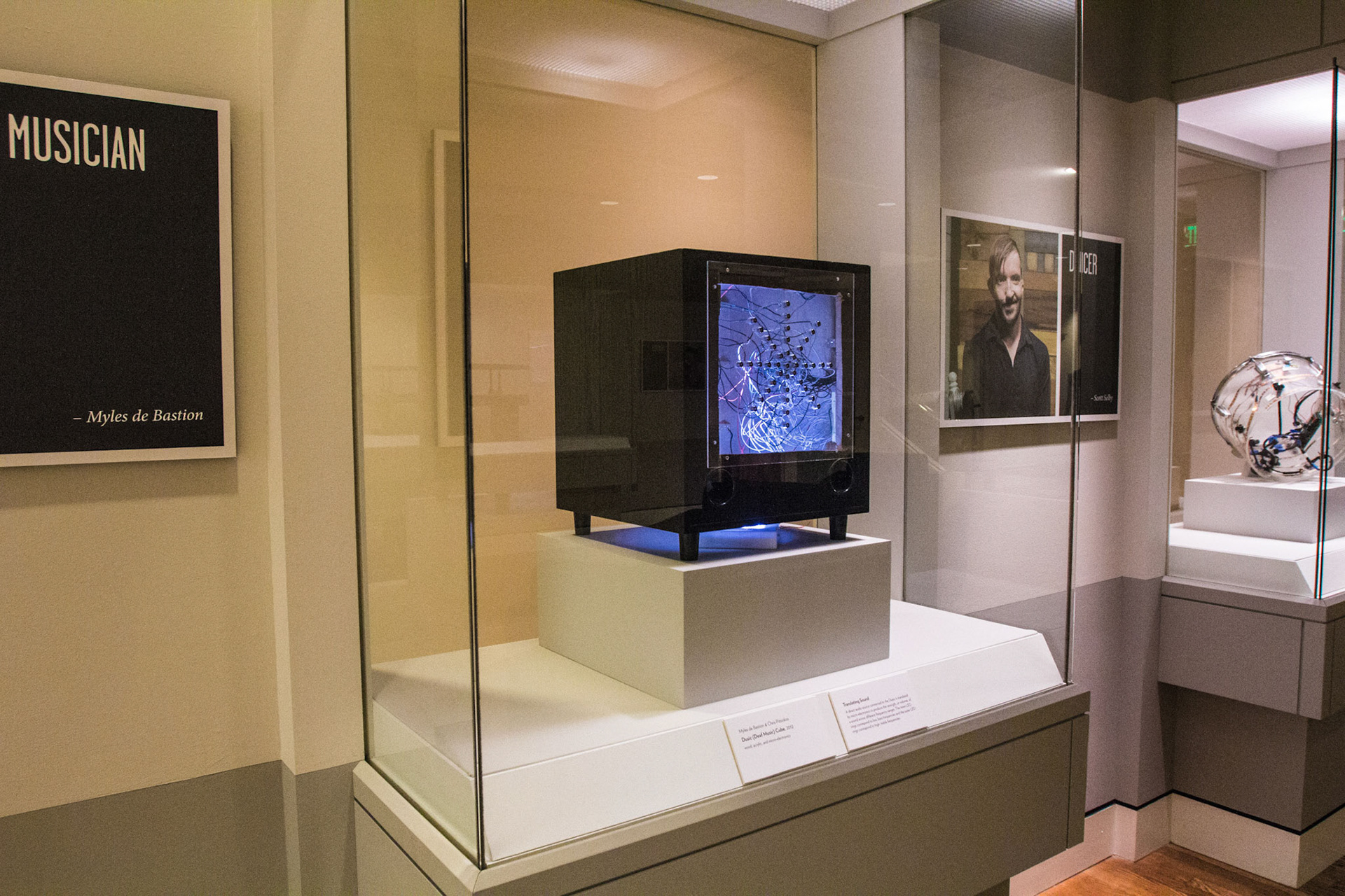 Gallery corner with the Dusic (Deaf Music) Cube glowing blue in its glass case, a black MUSICIAN panel credited to Myles de Bastion on the wall at left, a DANCER portrait of Scott Selby at right, and the clear drum visible in the case beyond
