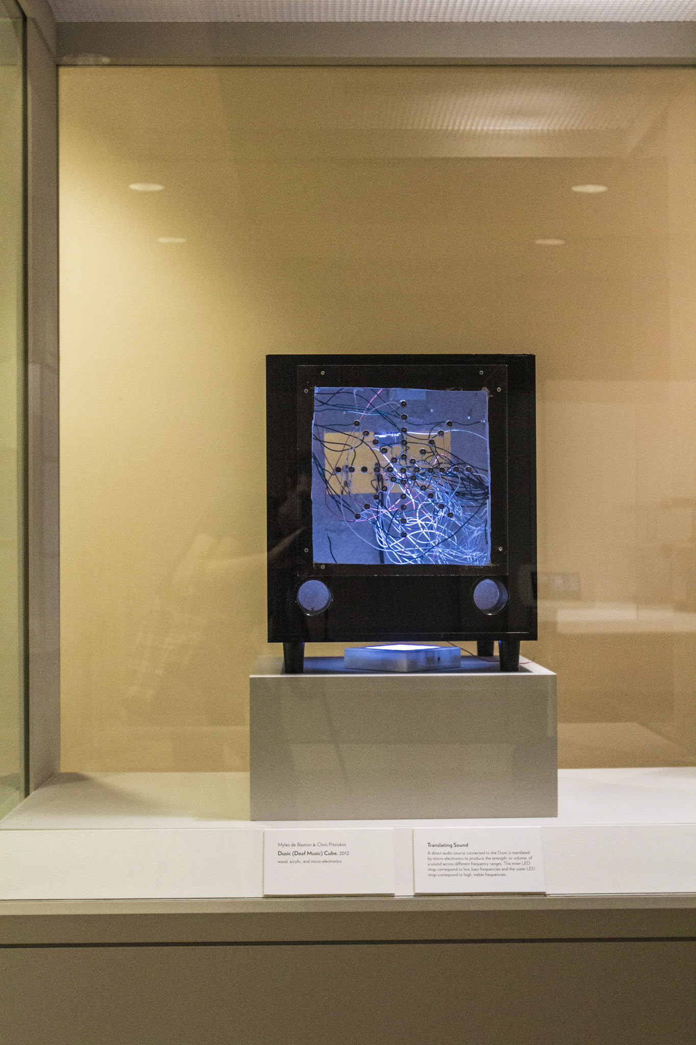 The Dusic (Deaf Music) Cube alone in its vitrine, a black wooden box whose front panel is a blue-lit tangle of white and pink wires studded with bolts, two speaker cones below and its label on the ledge