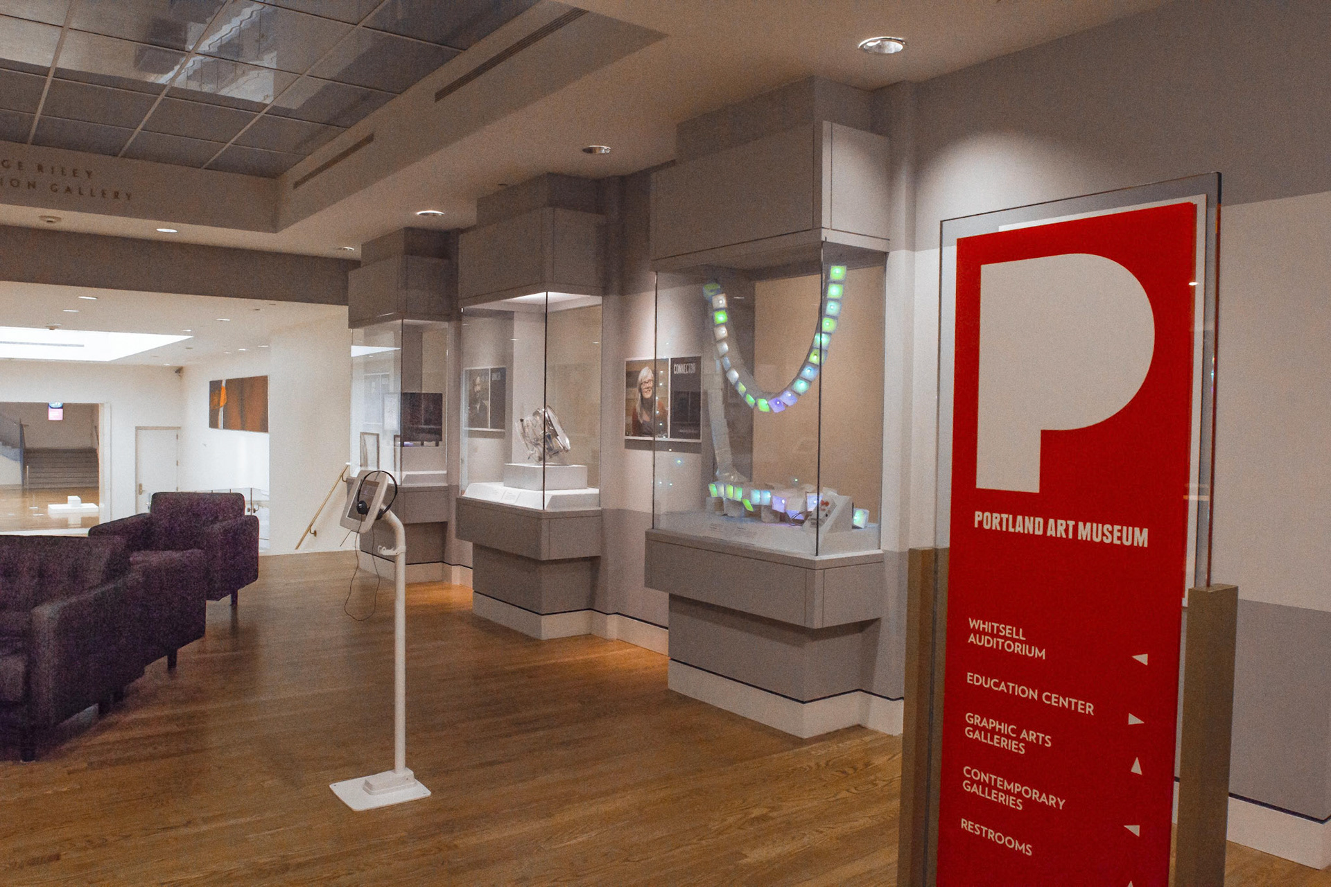 The red Portland Art Museum wayfinding sign in the foreground listing Whitsell Auditorium, Education Center and Graphic Arts Galleries, with the lit exhibition cases and a curving chain of green and white LED cubes down the gallery behind it