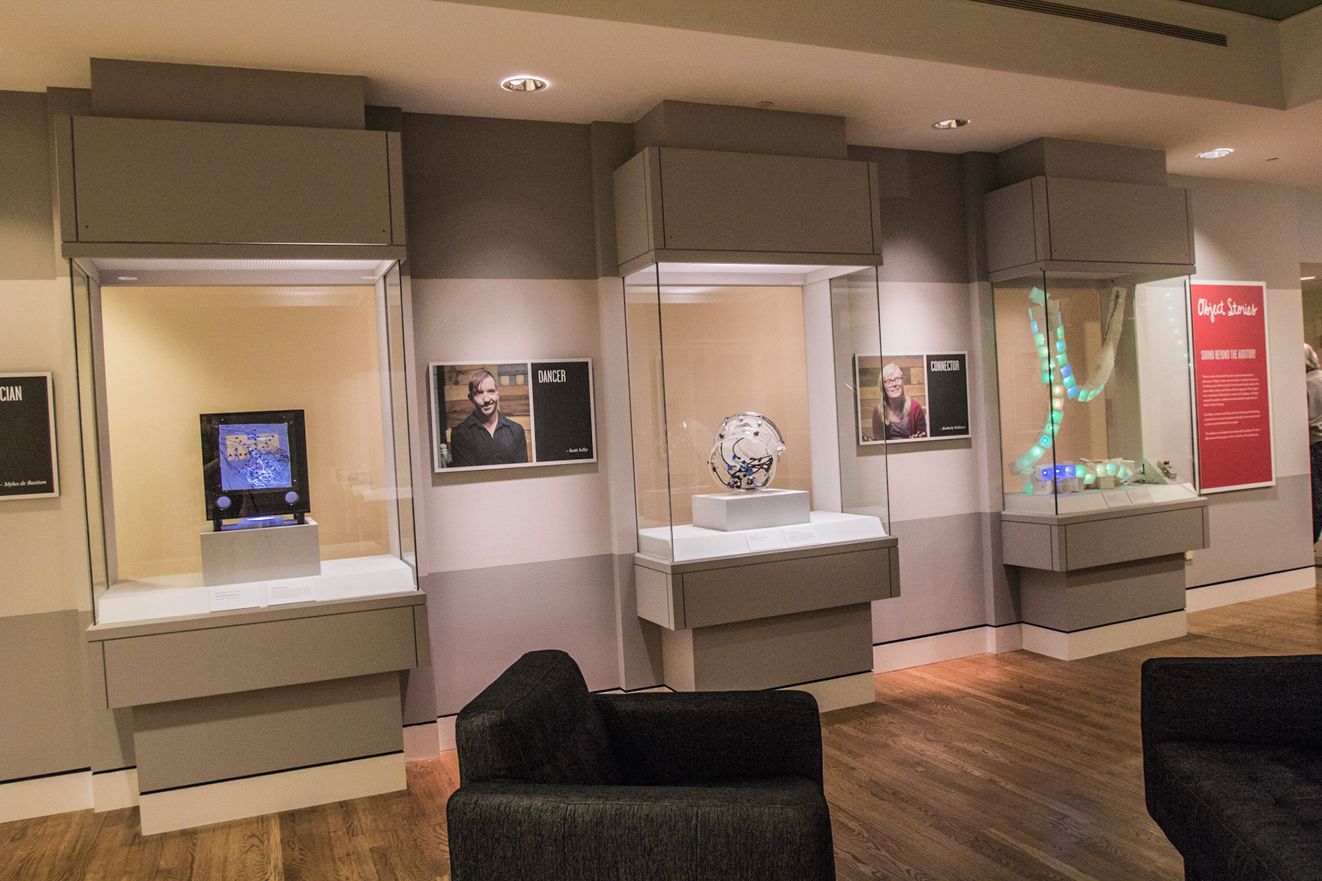 The row of three lit vitrines along the gallery wall holding the Dusic Cube, the clear drum and the curving LED cube chain, with DANCER and CONNECTOR portrait panels hung between them and the red Object Stories: Sound Beyond the Auditory panel at the far right