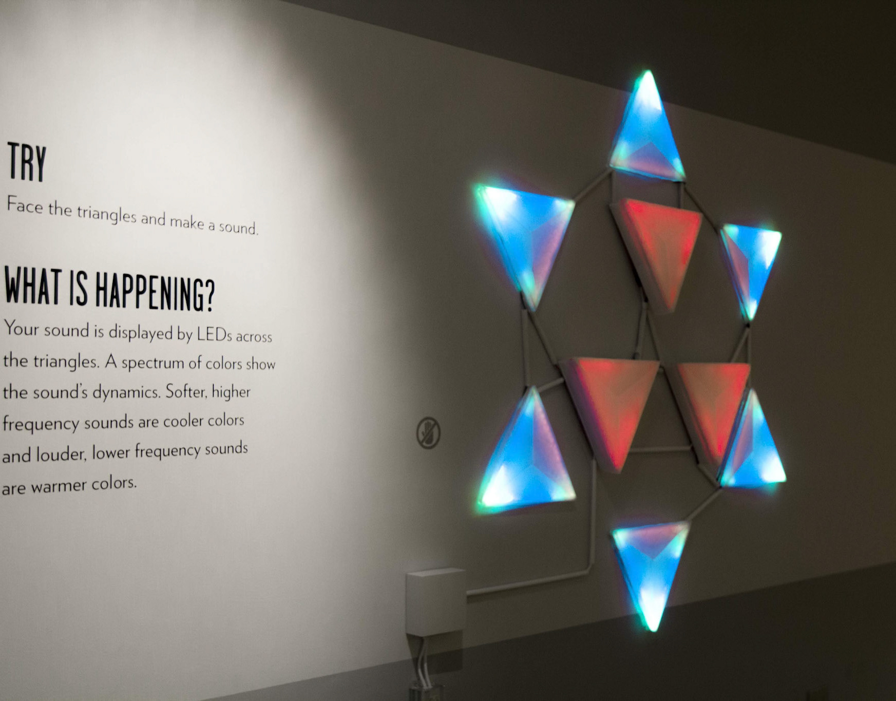 Illuminated triangular panels arranged as a star on a white gallery wall beside exhibition text