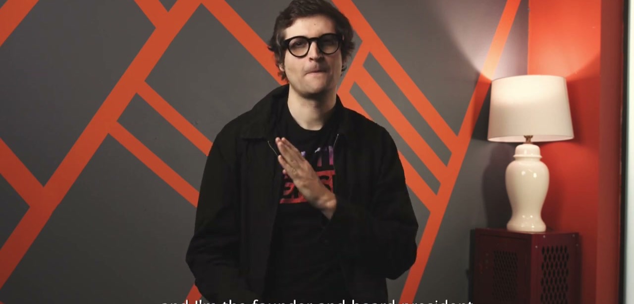Myles de Bastion signing to camera in front of an orange and gray geometric backdrop